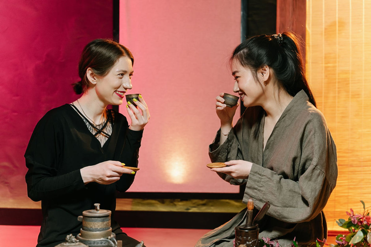 Two women smiling and drinking tea during a traditional tea ceremony indoors.